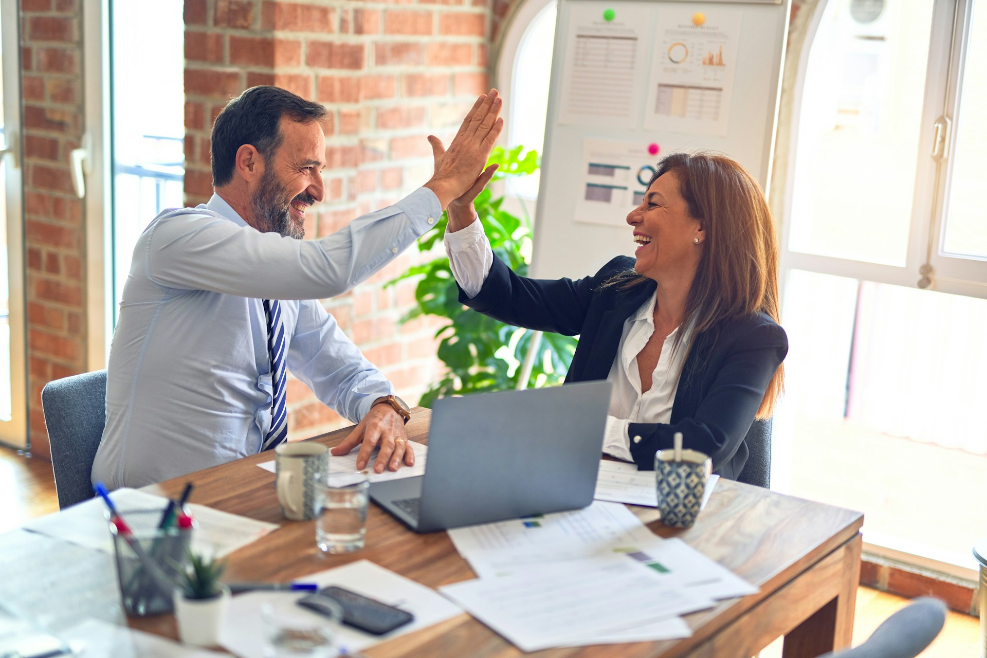 Two people sitting at a table with a laptop and scattered papers, high fiving